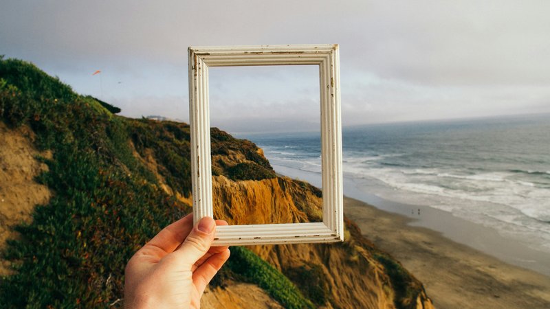 Hand houdt een leeg wit fotolijstje omhoog voor een kustlandschap met zee en klif, symbool voor het vastleggen van mooie momenten